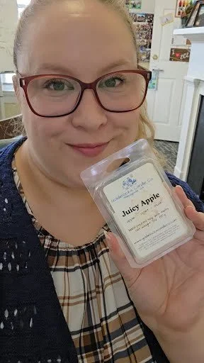 A smiling woman holding a Juicy Apple wax melt in clear packaging with a white label, indoors in a well-lit room.