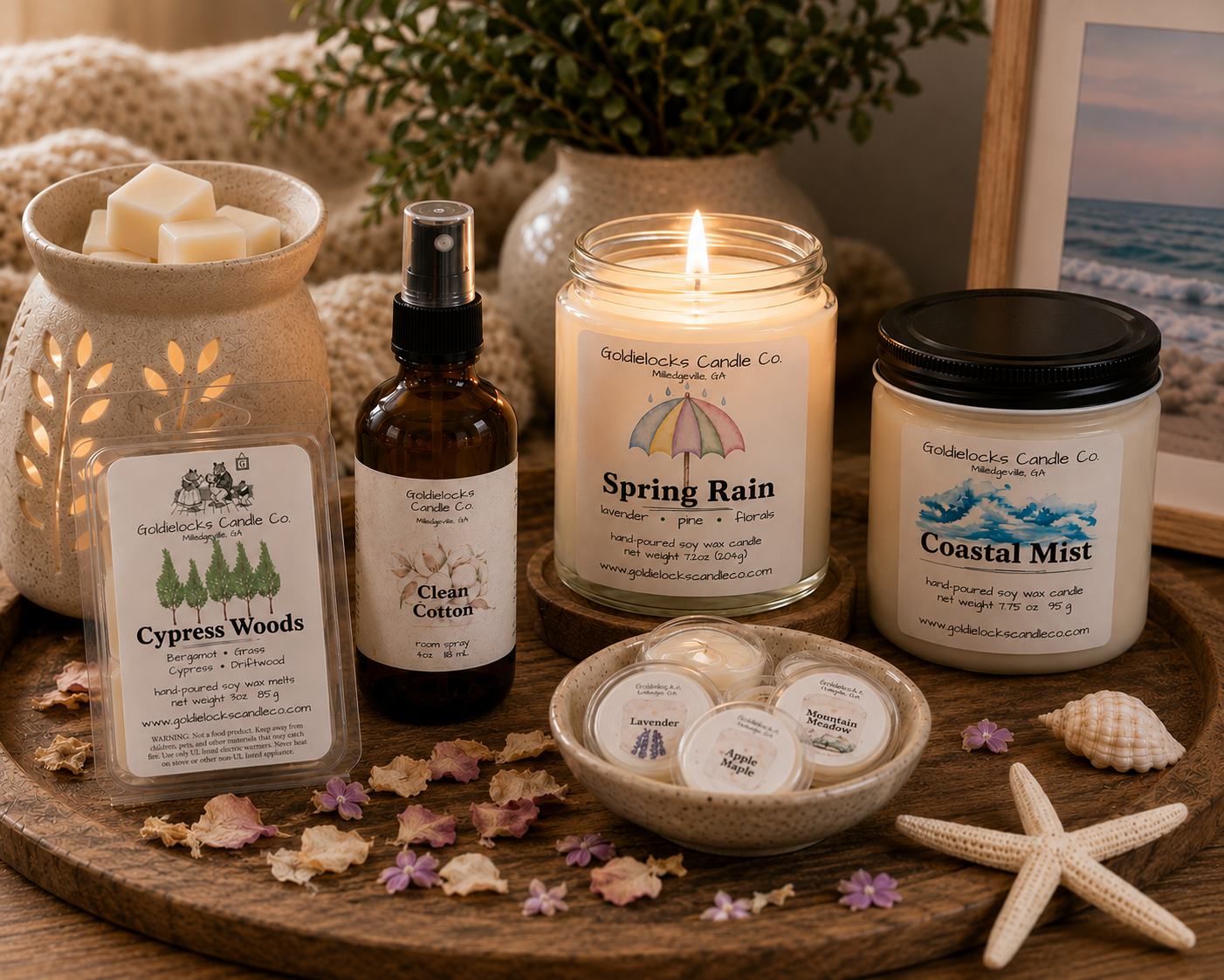 A collection of home decor and scented candles by Goldielocks Candle Co on a wooden tray, including a lit candle, a jar of Coastal Mist candle, a spray bottle labeled Clean Cotton, a small dish with scented wax melts, and decorations.