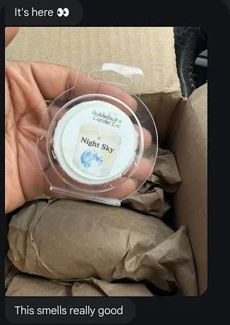 A person holding a small round container of a night sky-scented candle, labeled "Night Sky," inside a clear plastic case, with shredded packing paper surrounding it.