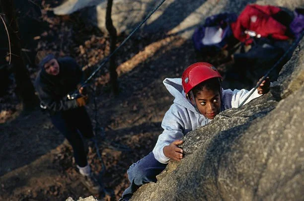 A young girl wearing a red helmet climbing a rock face outdoors, with another person in the background also climbing.
