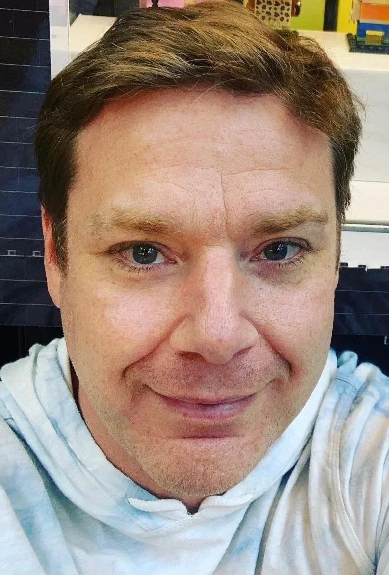 A close-up selfie of a smiling man with short brown hair and blue eyes, wearing a light-colored hoodie, in an indoor setting.