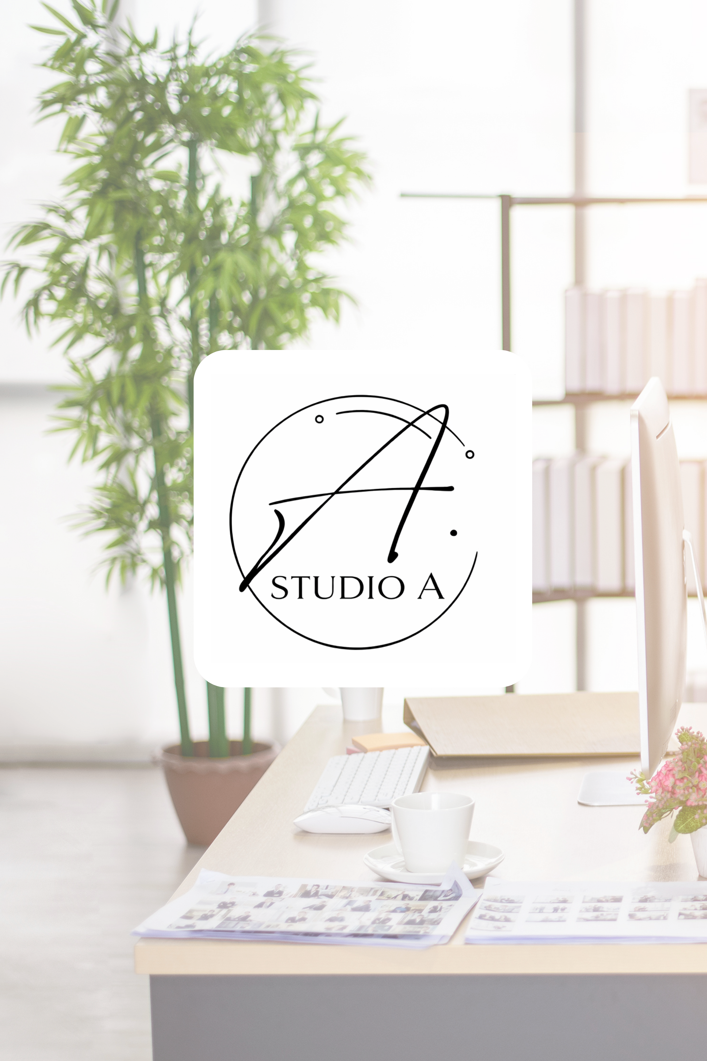 An office workspace with a computer monitor, keyboard, mouse, coffee mug, open photo album, and plant in the background. The computer screen displays a logo for Studio A, which features a large stylized letter A with decorative lines and circles.