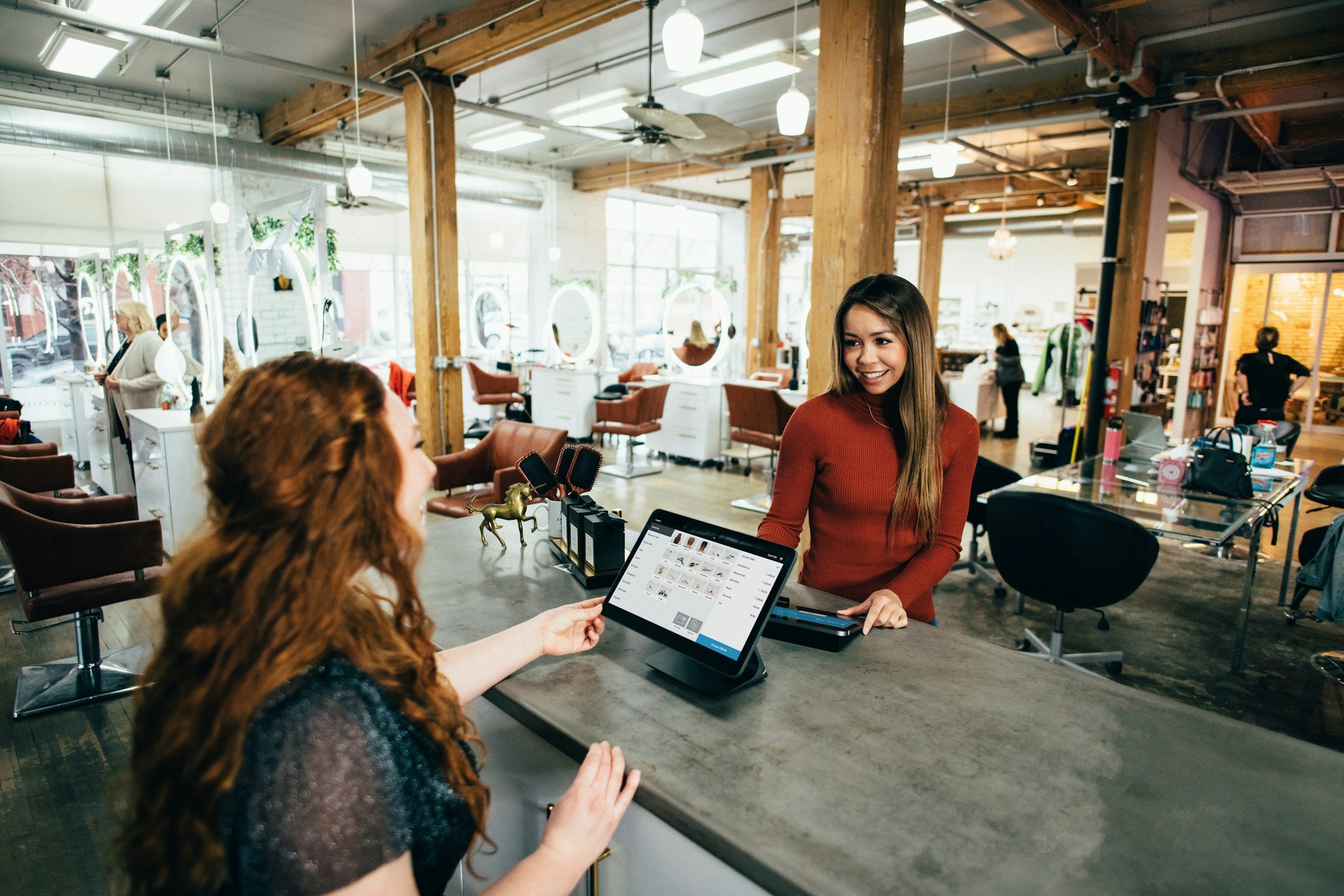A woman with long brown hair in a red top is smiling at a waitress with long curly red hair who is handing her a card at a salon reception desk. The salon has large windows and wooden beams, with several salon chairs and mirrors visible in the background.
