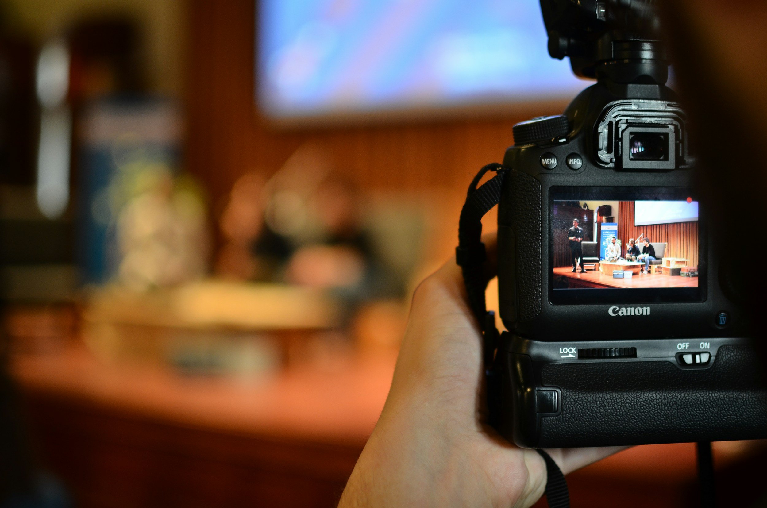 Photograph of a Canon camera recording a panel of speakers on a stage with wooden wall background.