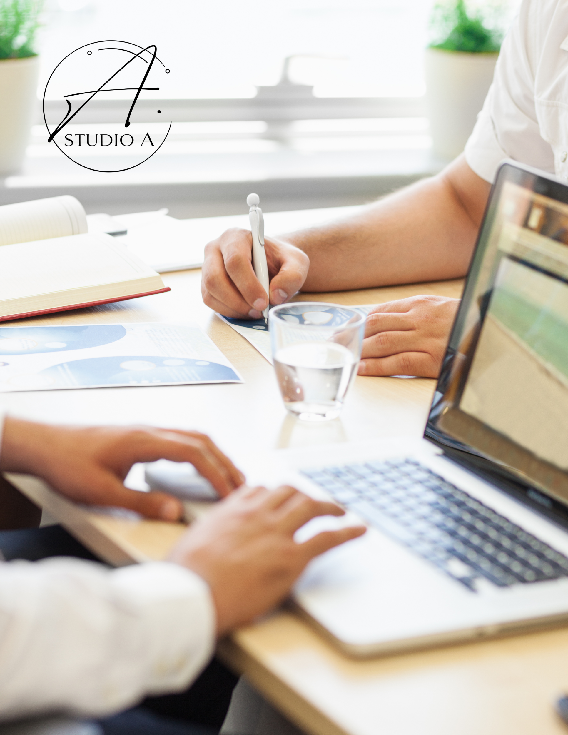 People working at a desk with a laptop, documents, a glass of water, and a person taking notes.