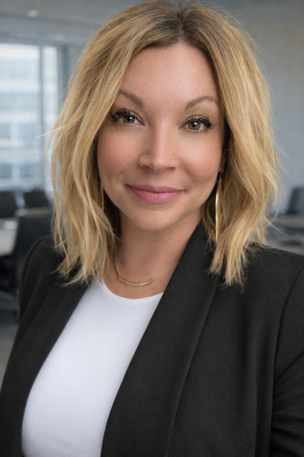A woman with shoulder-length blonde hair and fair skin, wearing a black blazer over a white shirt, smiling in an office setting.