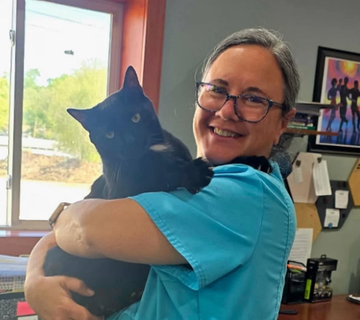 A woman with glasses and a blue shirt holding a black cat inside a room near a window, smiling at the camera.