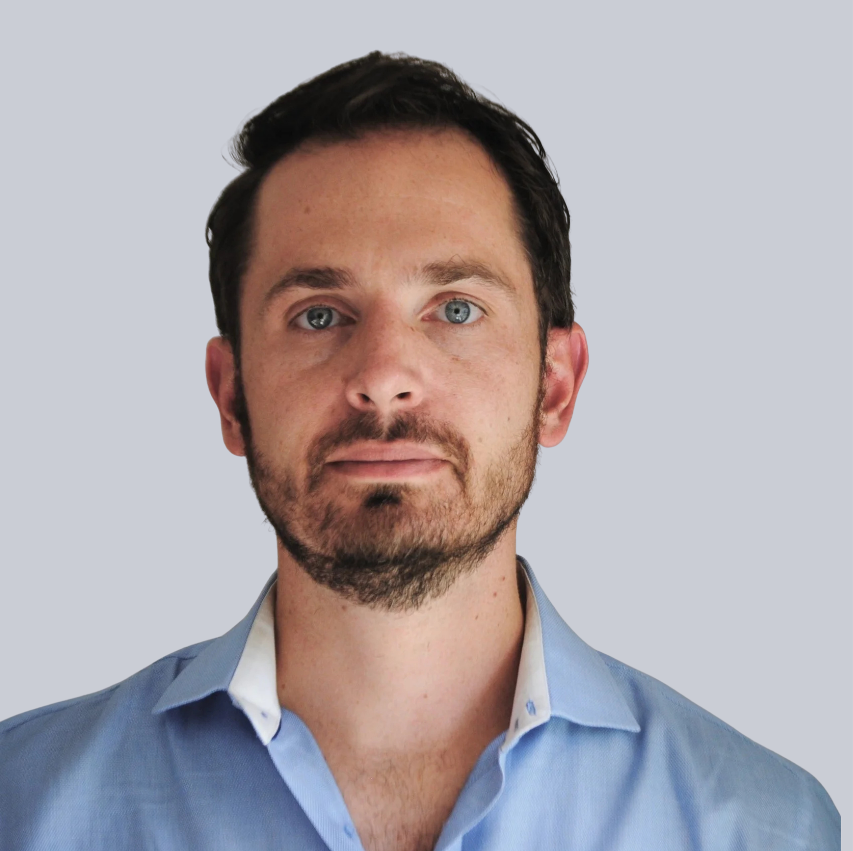 Close-up of a man's face showing his light blue eyes, dark hair, and beard, wearing a light blue collared shirt against a plain light background.