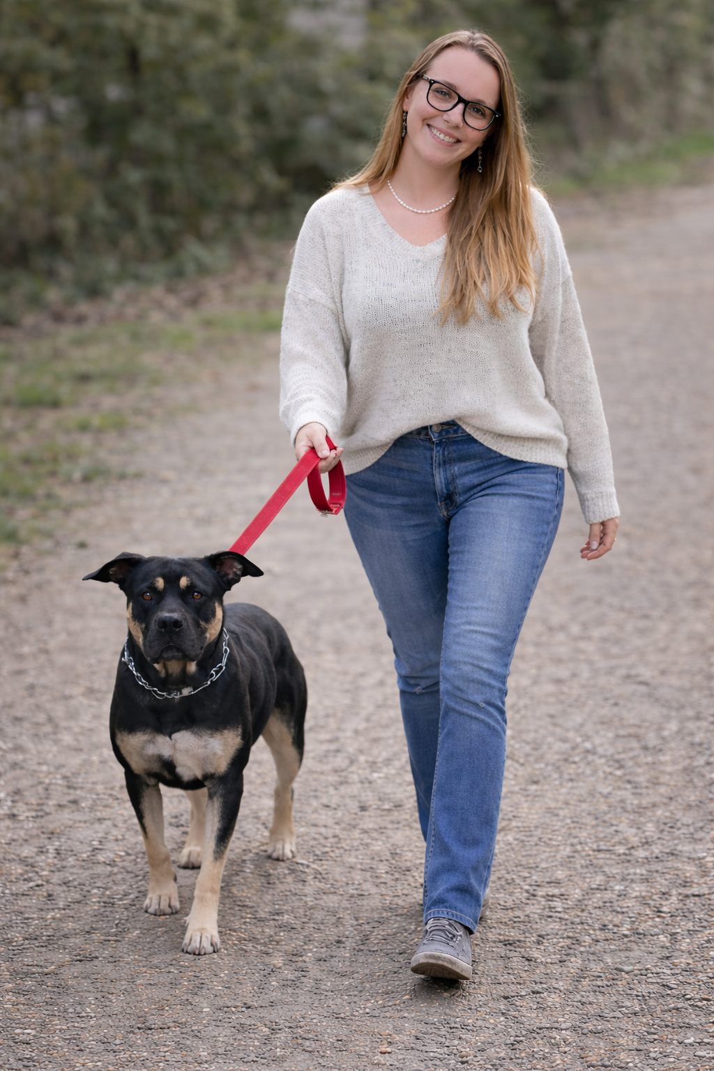 A smiling woman with glasses, wearing a light-colored sweater and jeans, walking a black and tan dog on a red leash outdoors on a dirt path.