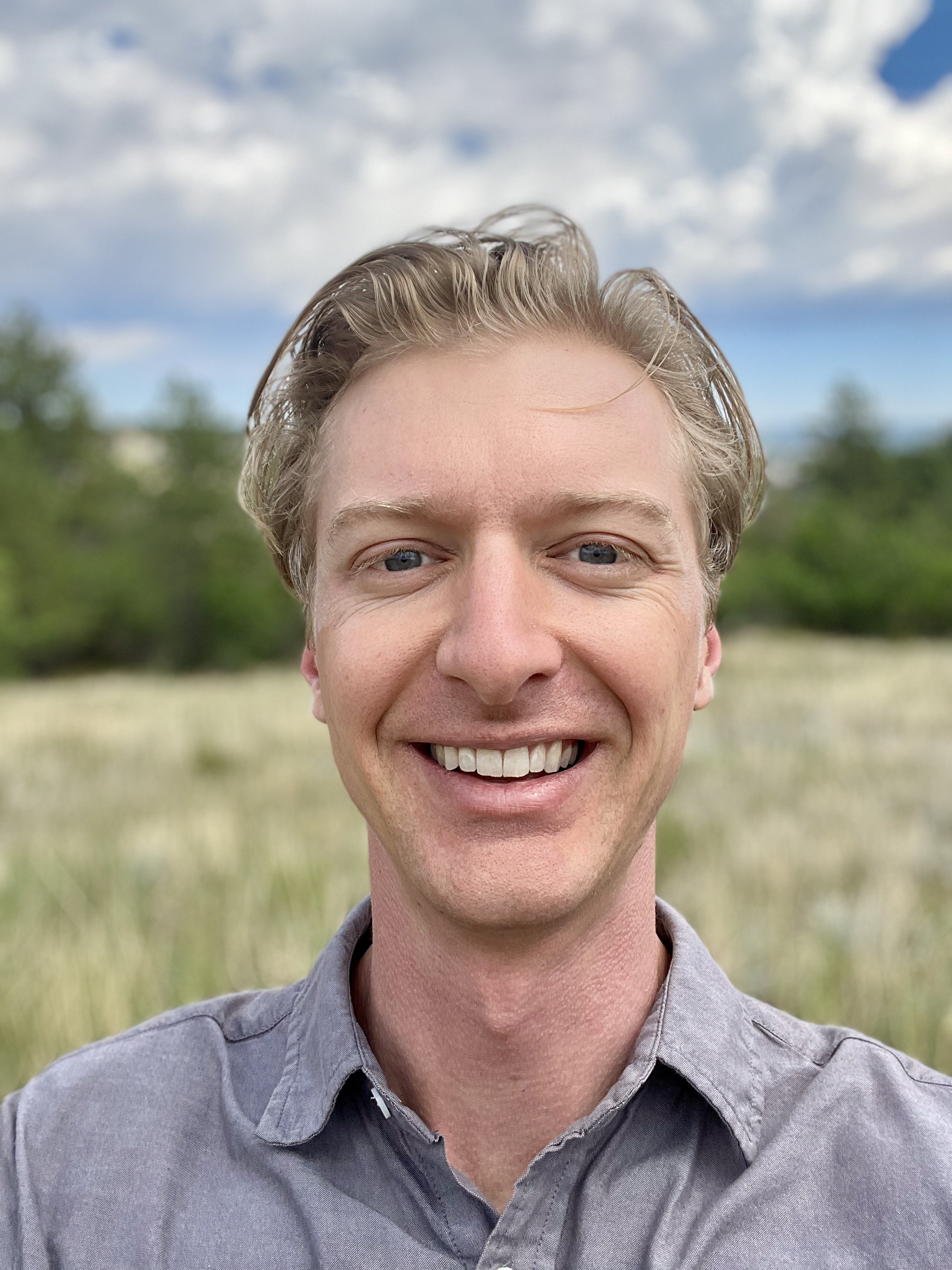 Close-up of a smiling man with blonde hair, blue eyes, wearing a gray button-up shirt, outdoors with a blurry green field and blue sky with clouds in the background.