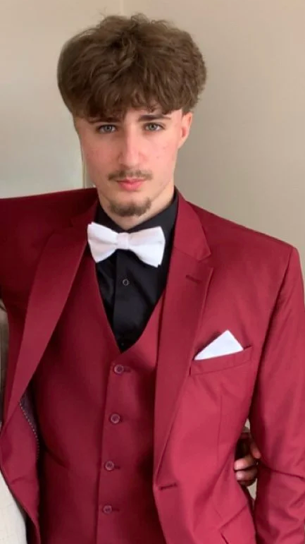 Young man with curly brown hair, mustache, and beard wearing a red suit jacket, matching vest, black dress shirt, white bow tie, and pocket square.