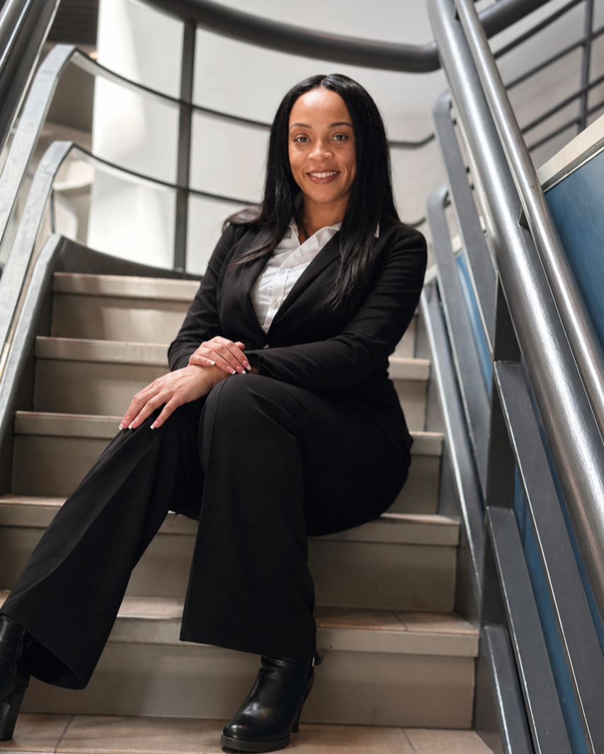 A woman in a black business suit with a white shirt, sitting on a staircase with metal handrails.