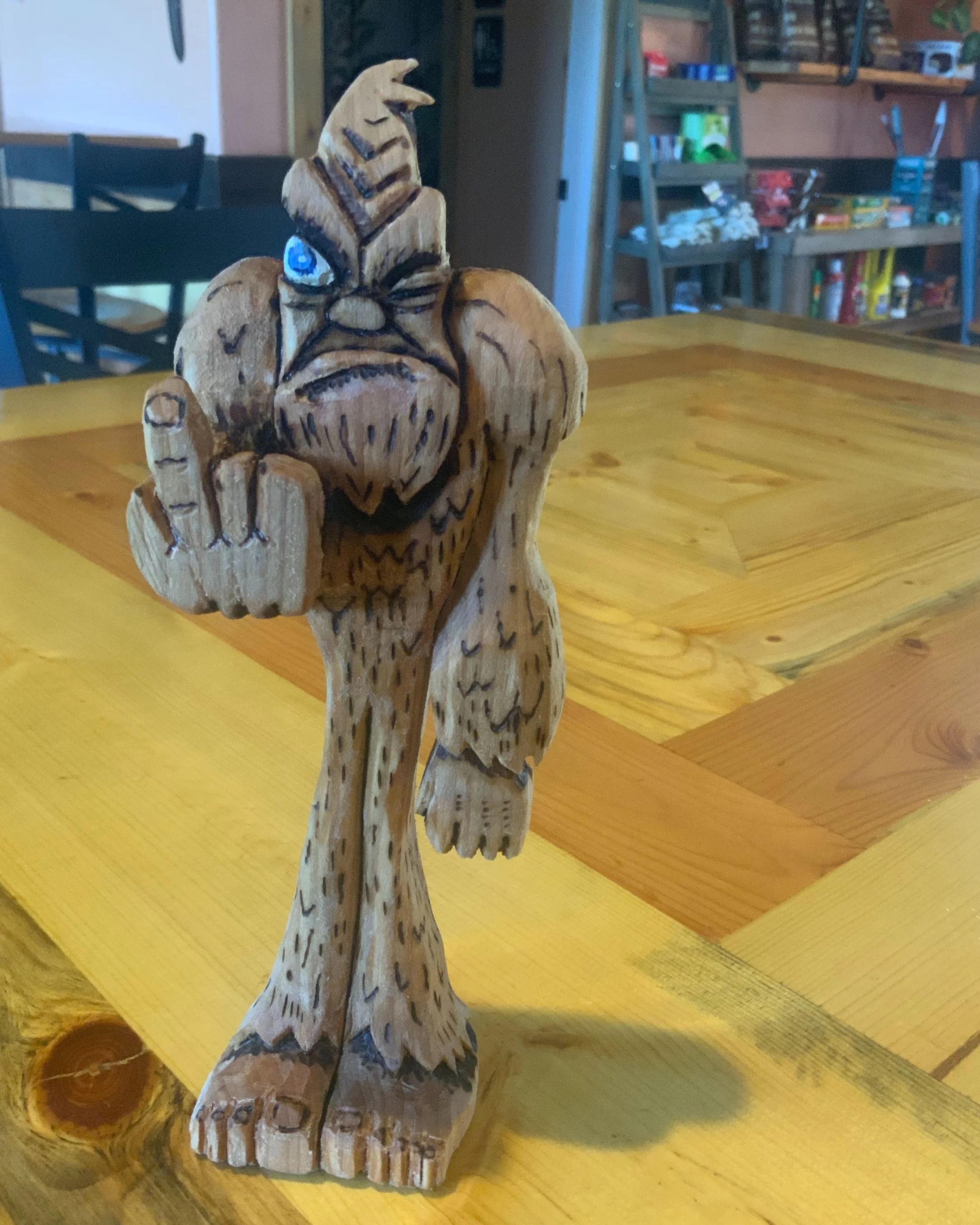 A wooden figurine of a bird with a beak, wings, and tail, carved from a single piece of wood, sitting on a wooden surface. The bird has a blue and white eye painted on its head.