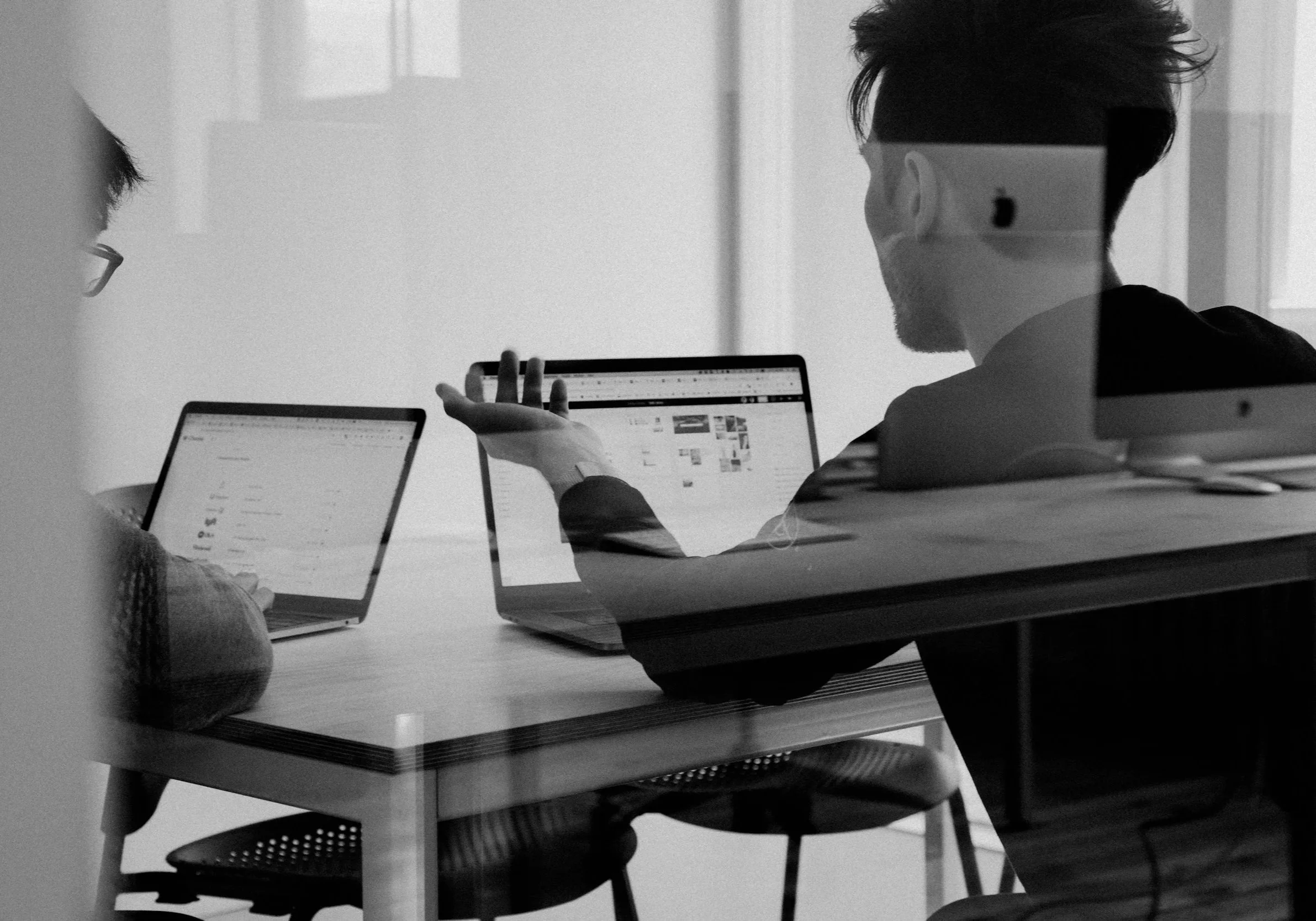 A black-and-white photo of three people sitting at a table, working on laptops. One person is wearing glasses and the other two have masks on. The scene appears to be in an office or meeting room.