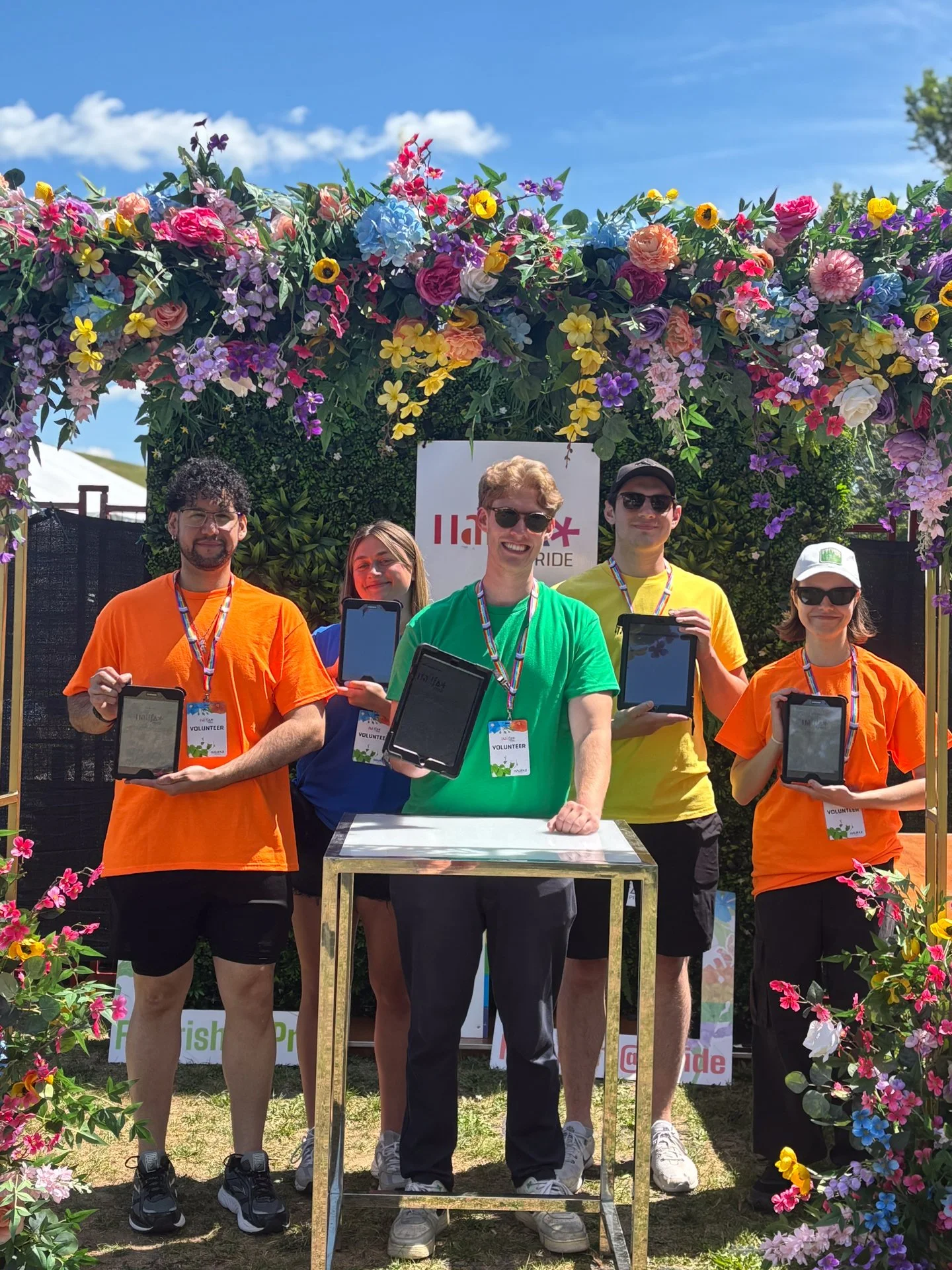Group of five volunteers standing under an arch of colorful flowers at an outdoor event, all holding tablets, with a sign behind them that says "Flagship Pride."