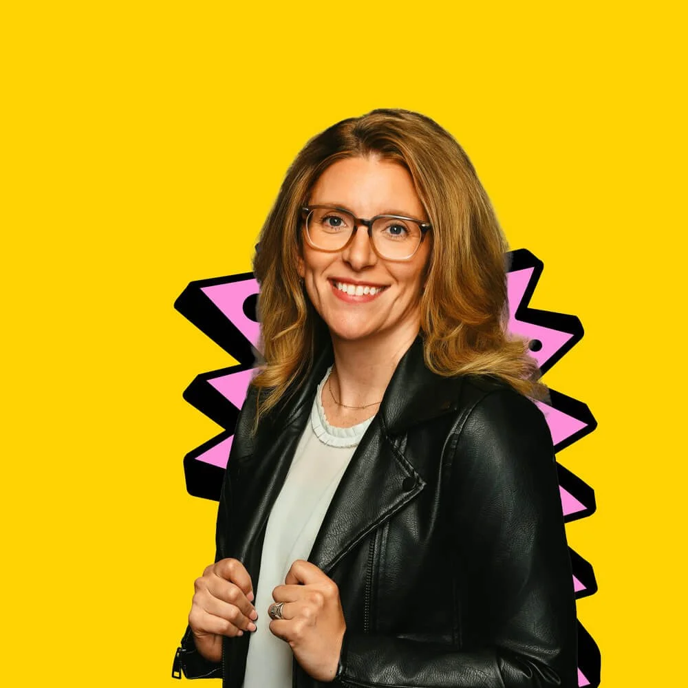 A woman with shoulder-length blonde hair, glasses, and a black leather jacket smiling against a vibrant yellow background with a pink and black abstract design.
