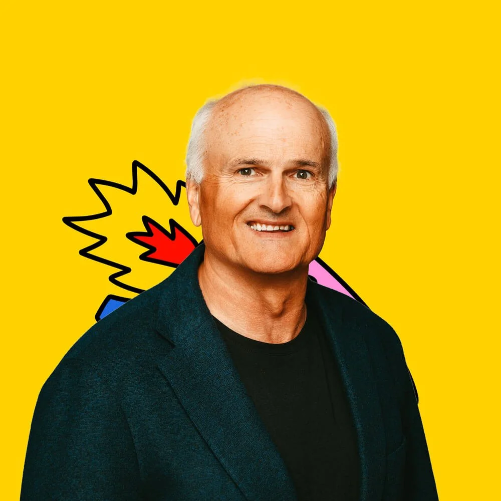 A smiling middle-aged man with gray hair, wearing a dark blazer and black shirt, standing in front of a yellow background with a cartoon explosion graphic and pink, blue, and red elements.
