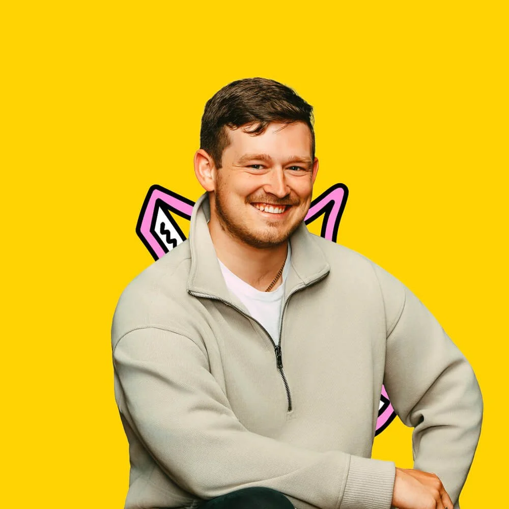 A smiling man with short brown hair and a beard, wearing a light gray zip-up jacket over a white shirt, poses against a bright yellow background with a pink and black cartoon style cat illustration behind him.