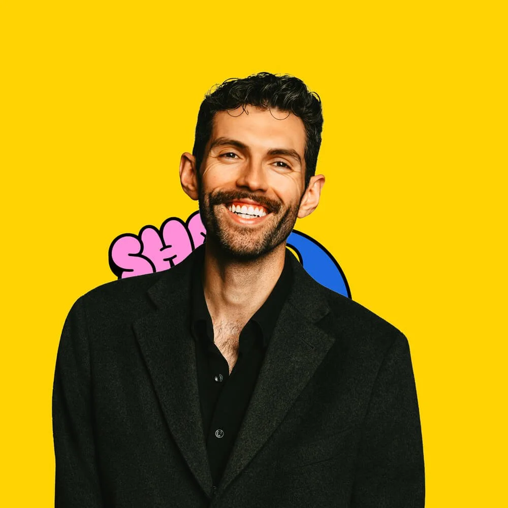 A man with dark curly hair and a beard, smiling, wearing a black jacket against a yellow background with partial pink and blue text.
