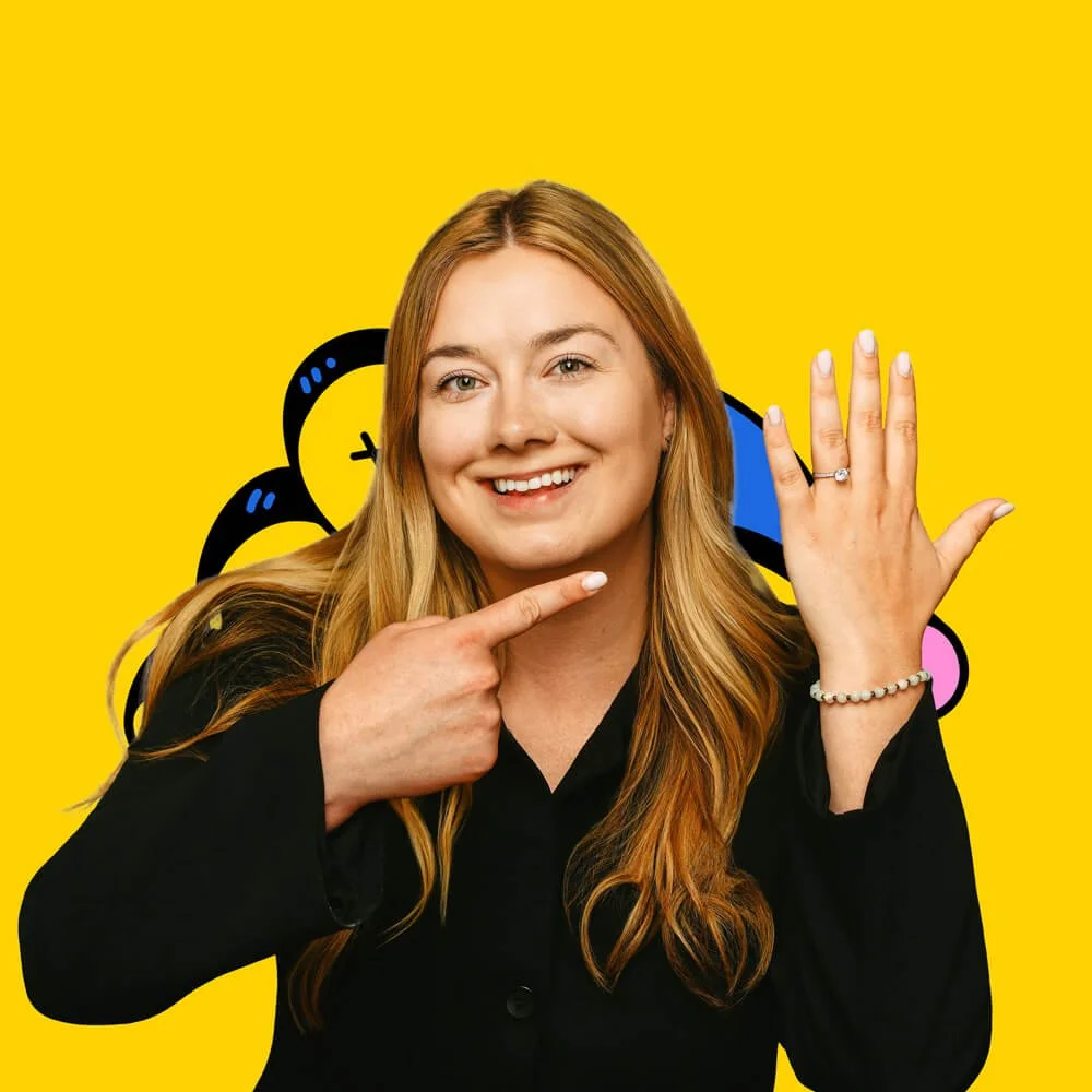 A smiling woman with long red hair pointing at her left hand which displays a ring on her finger. She is wearing a black top and a pearl bracelet, set against a yellow background with blue, black, and pink graphic elements.