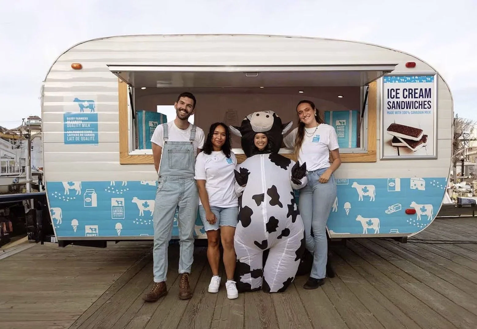 Four people standing with a person in a cow costume in front of an ice cream truck. The truck has signs for ice cream sandwiches and dairy products.