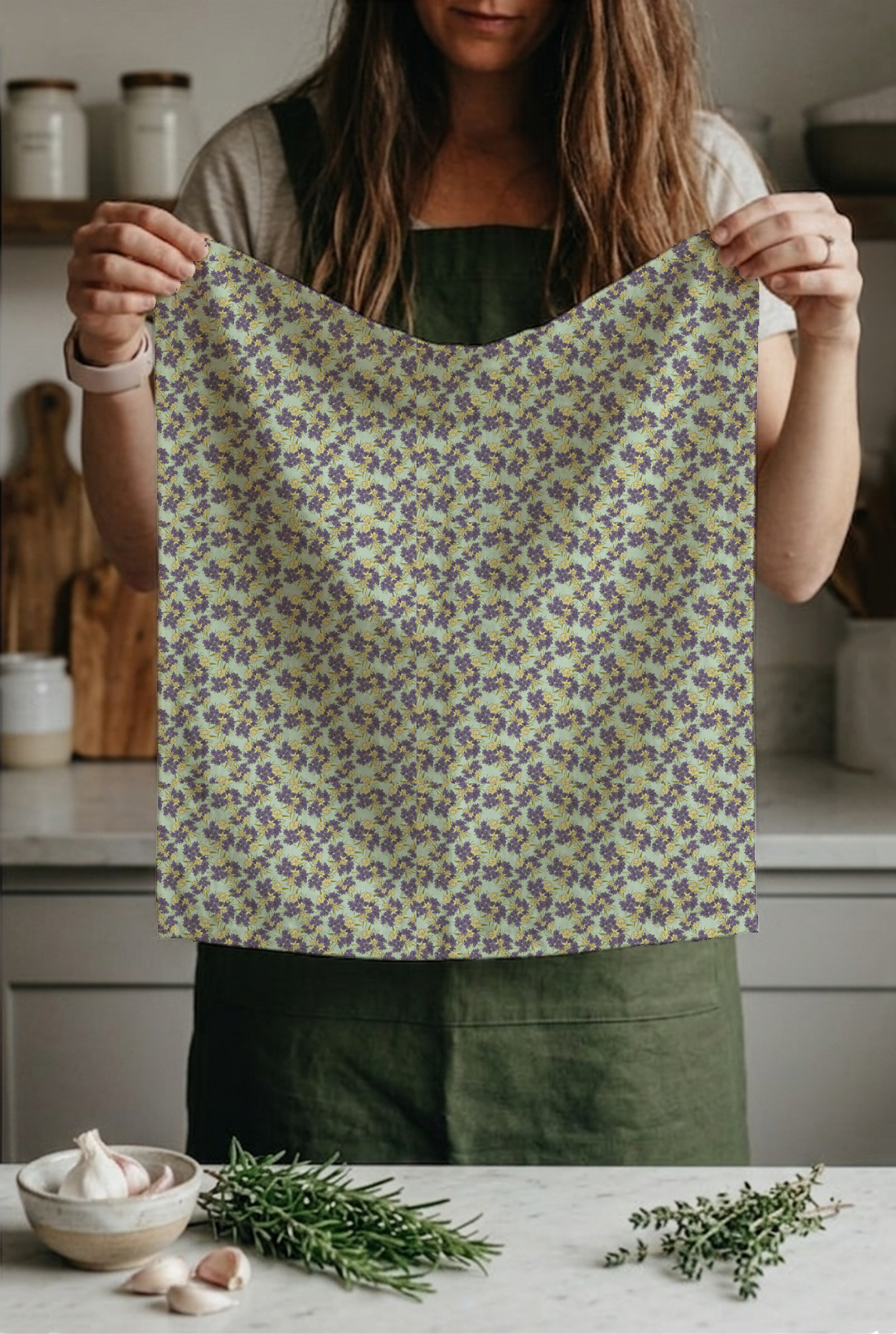A woman in a gray shirt and apron holds a purple and yellow floral cloth in a kitchen with garlic and fresh herbs on a white countertop.