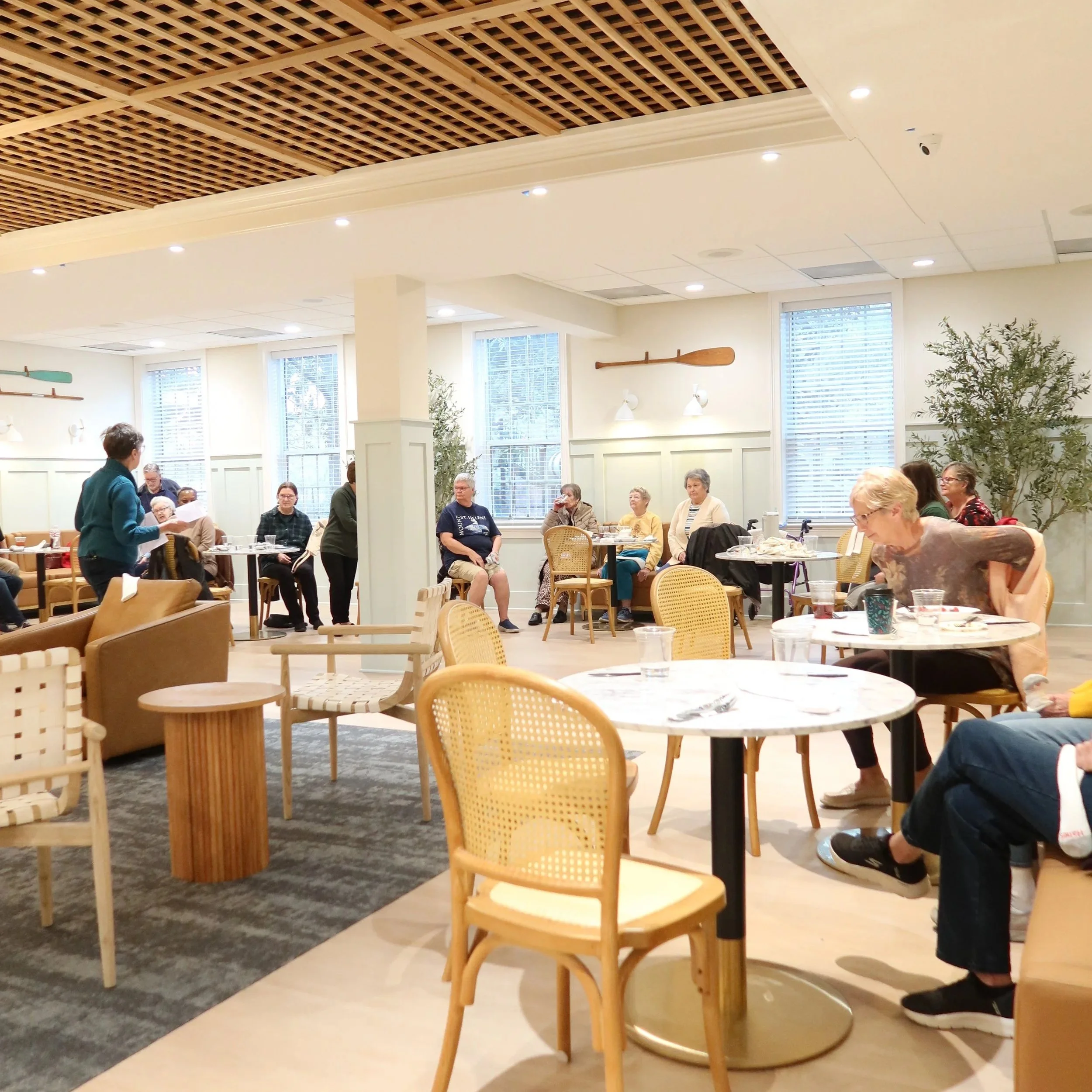 People sitting and standing in a bright, modern room with large windows, white walls, and wooden furniture, some engaged in conversation and others having food or drinks.