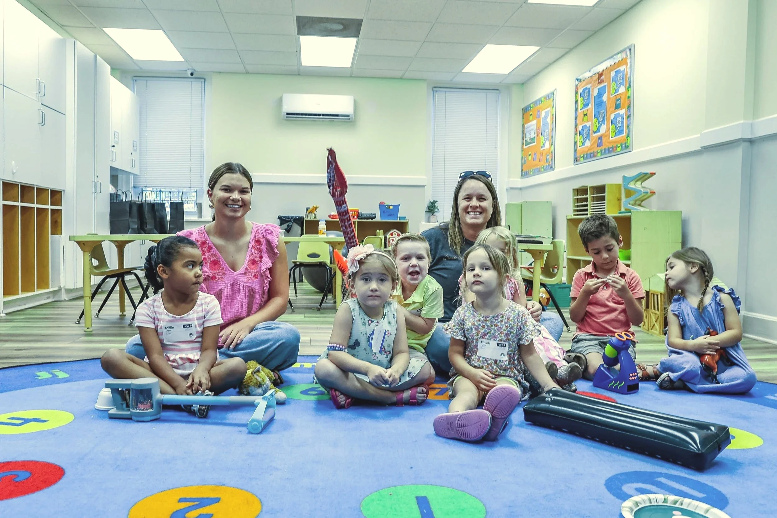 A group of children and two women sitting on a colorful alphabet-themed rug in a classroom. The children have name tags and are holding or surrounded by toys and learning tools. The classroom has bright lighting, educational posters, and storage units.