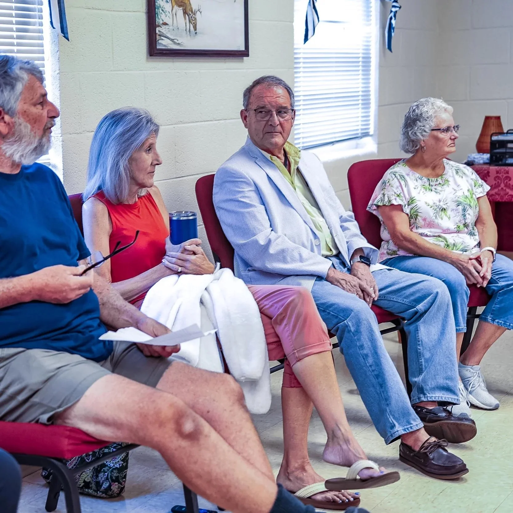 A group of elderly people sitting on red chairs in a room with beige walls and windows with blinds. The people are engaged, some listening attentively. There is a painting of deer on the wall behind them.