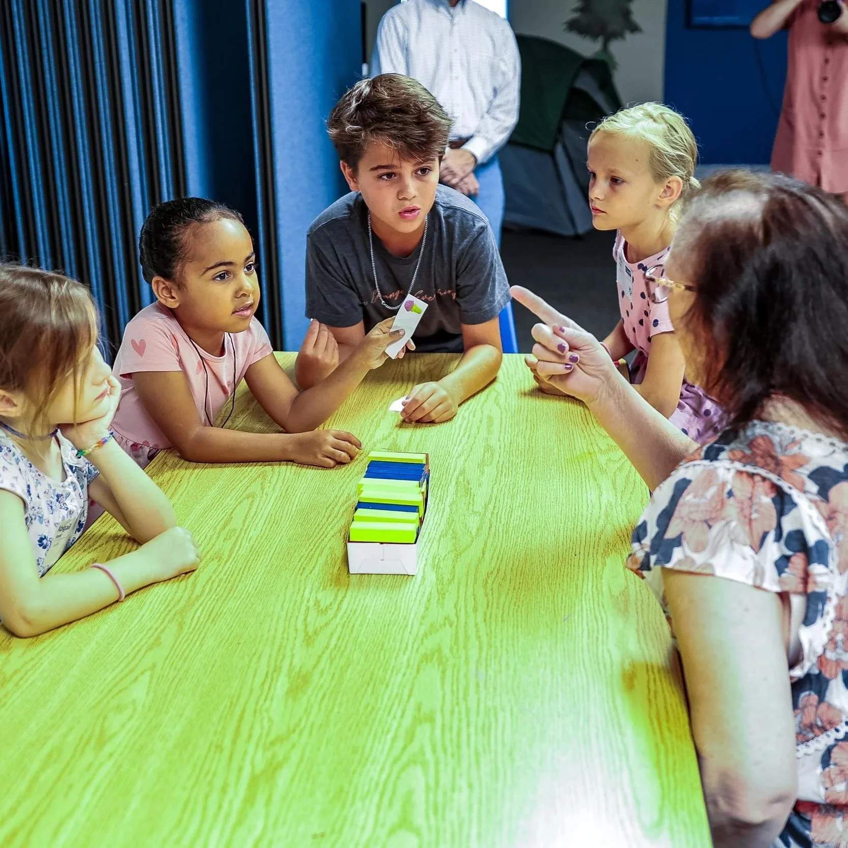 Children sitting around a green table playing a card game, with a stack of cards in the center of the table, inside a room with blue walls.