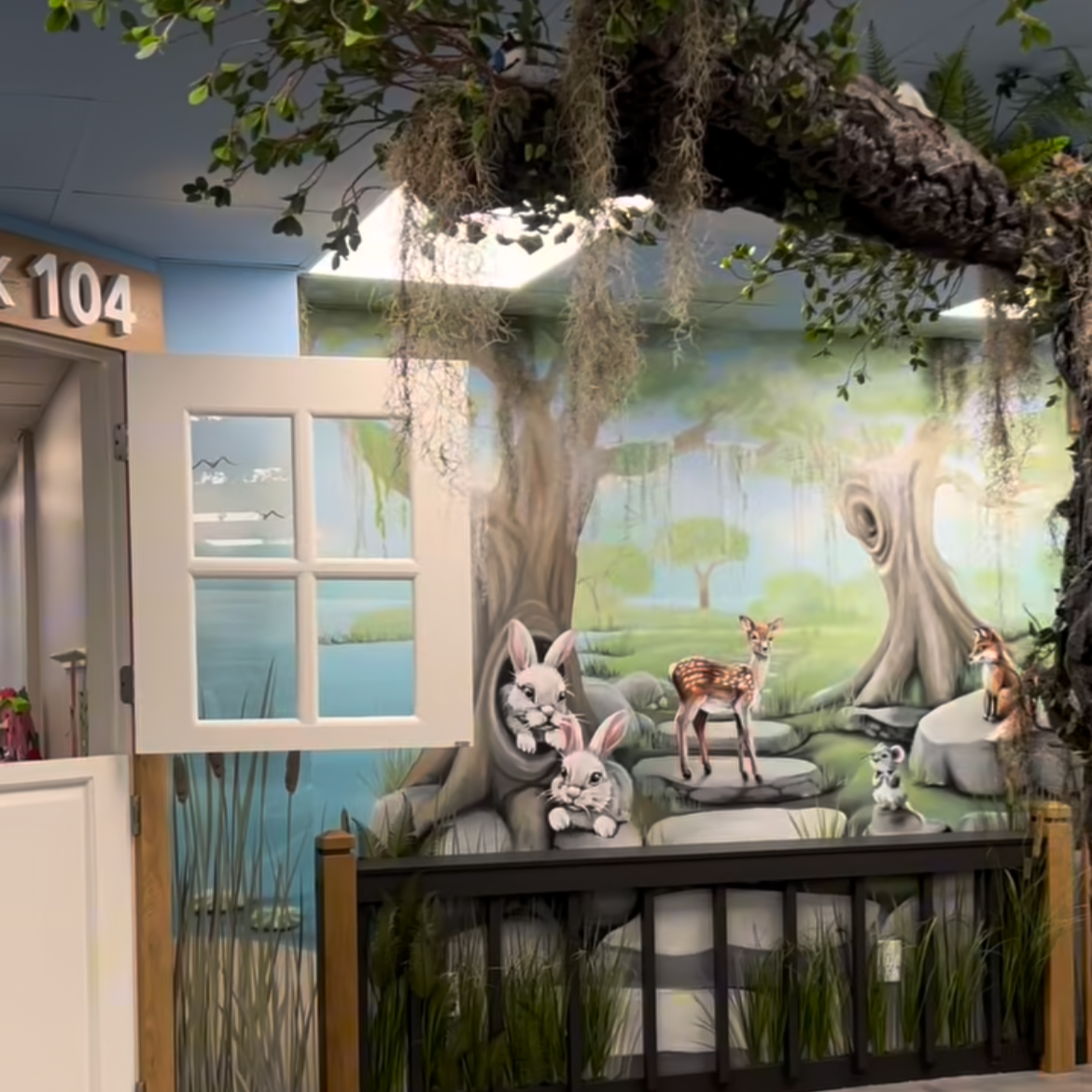 Decorative mural in a children's area depicting a forest scene with a rabbit and a deer surrounded by trees, grass, and water features.
