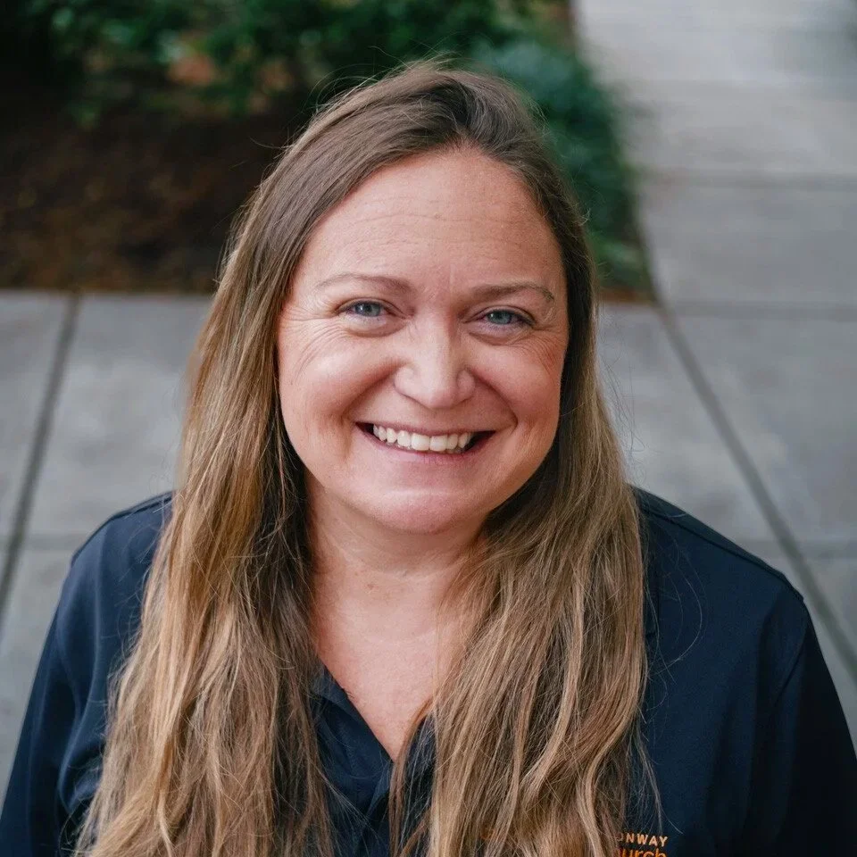 A smiling woman with long light brown hair, wearing a dark blue shirt, outdoors with a sidewalk and green bushes in the background.