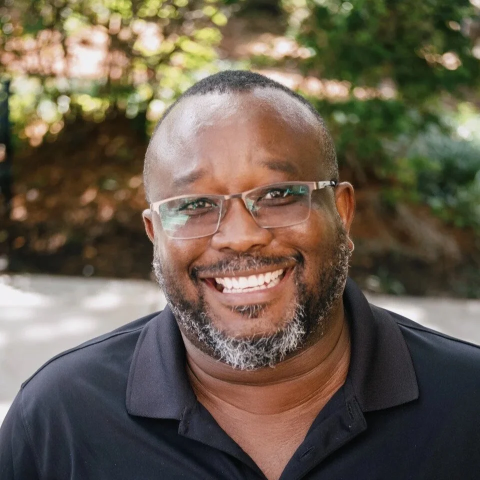 A smiling man wearing glasses and a black polo shirt outdoors with greenery and trees in the background.