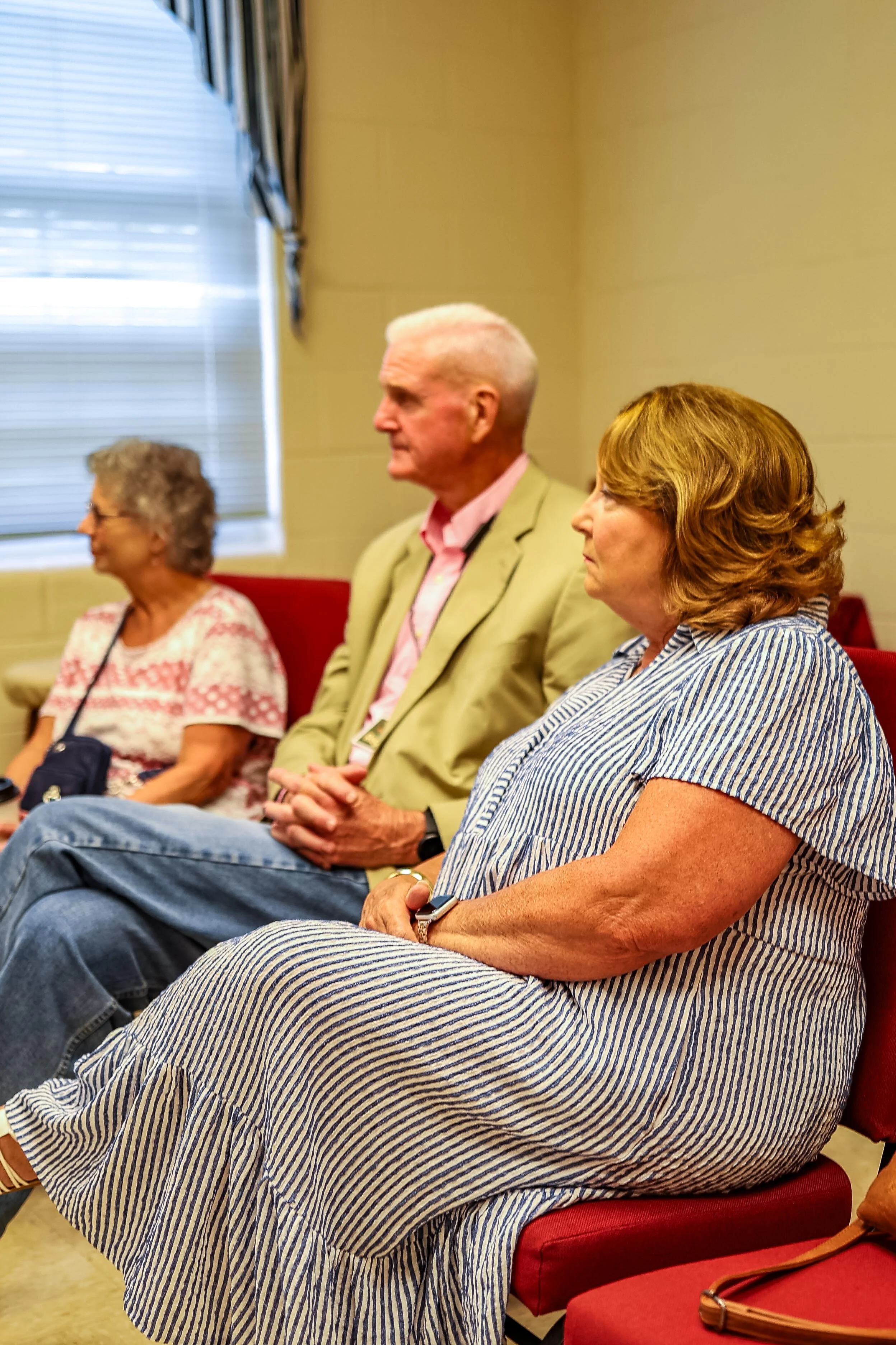 Three elderly individuals sitting in a row at a meeting or event, with two women on either side and a man in the middle, all facing to the right.