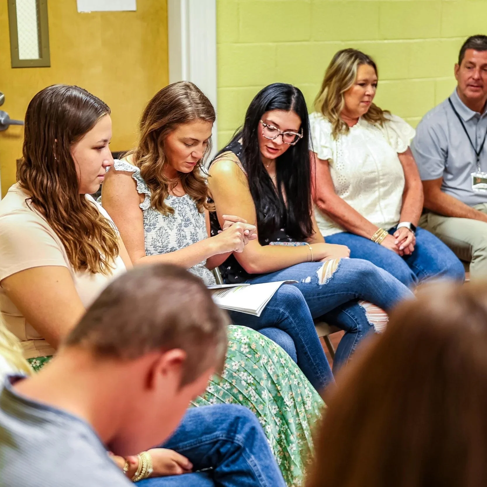 A group of people sitting in a row, participating in an indoor event or meeting, with some reading and others listening.