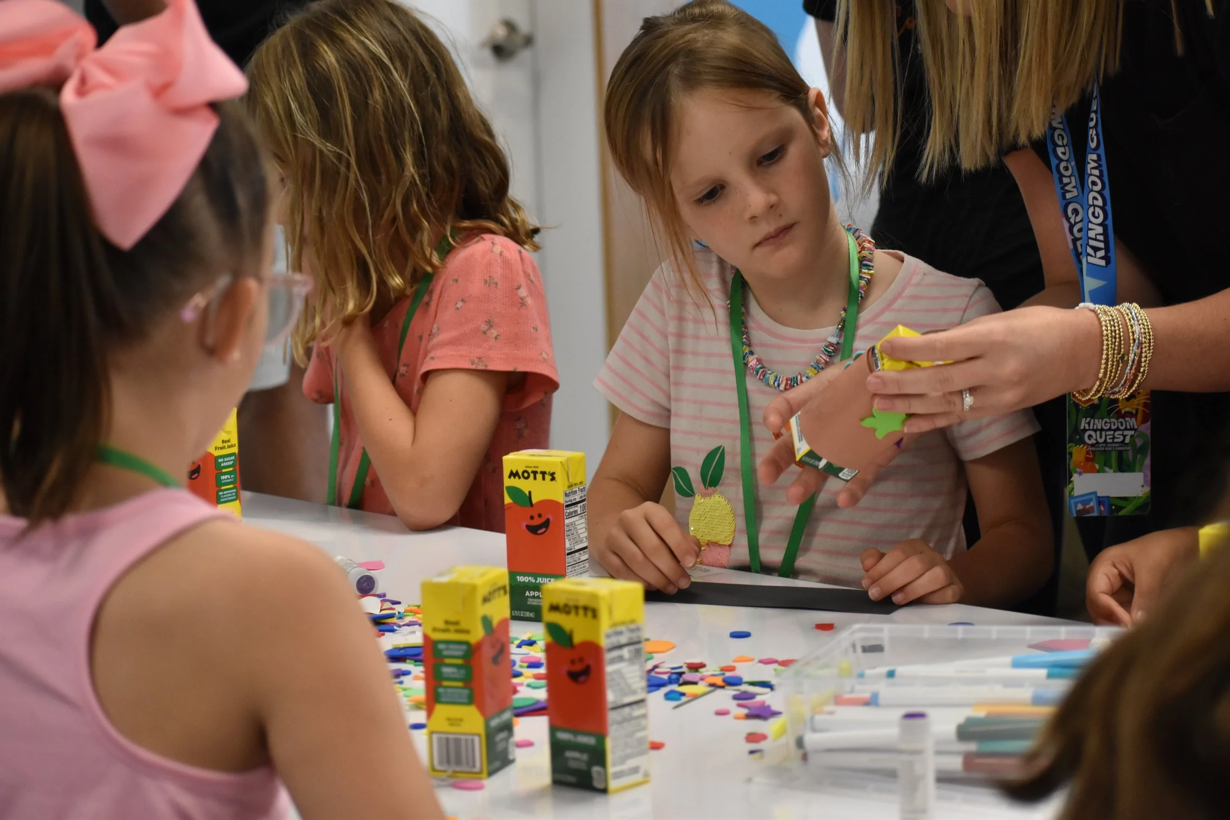 A group of children and two women sitting on a colorful alphabet-themed rug in a classroom. The children have name tags and are holding or surrounded by toys and learning tools. The classroom has bright lighting, educational posters, and storage units.