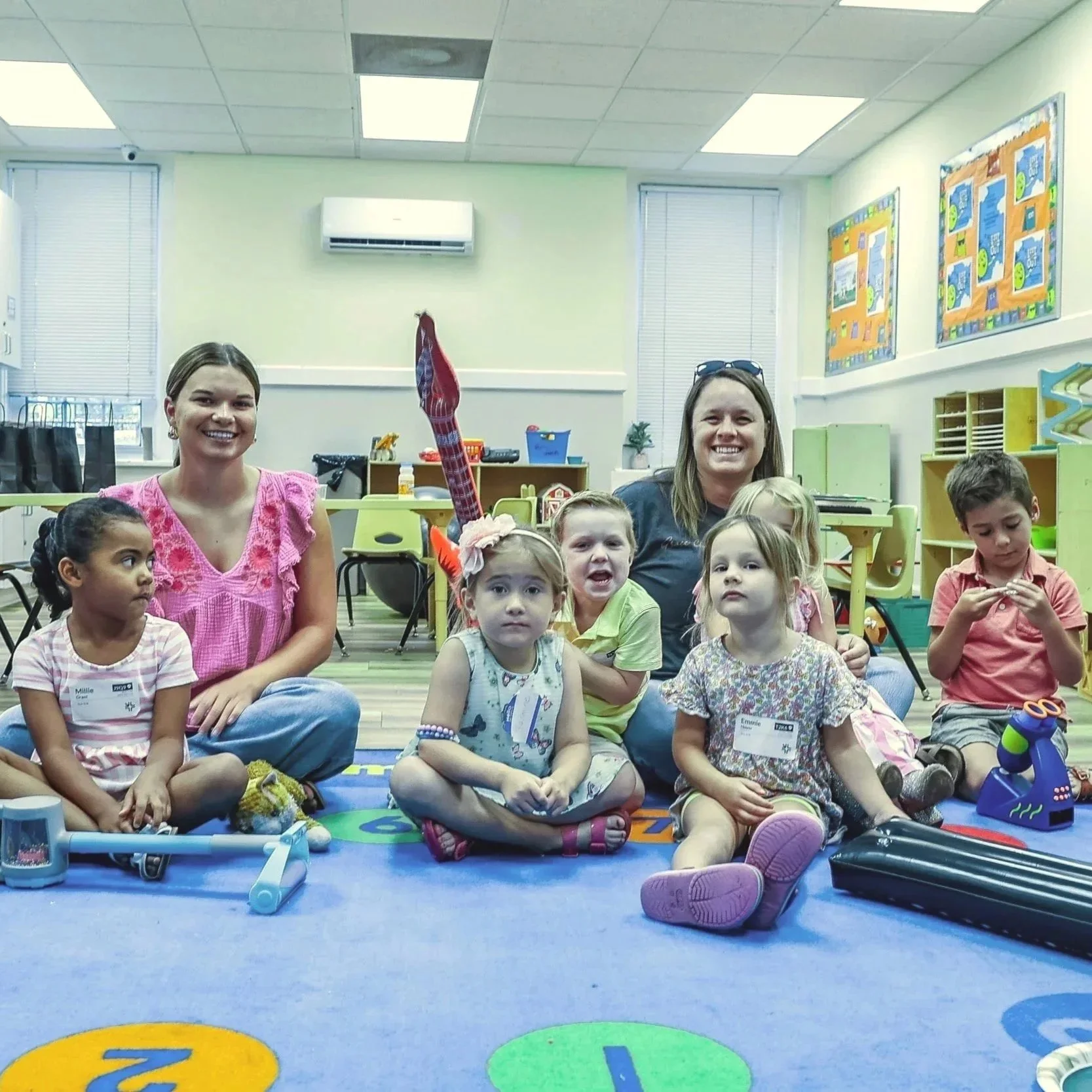 Group of children and two adult women sitting on a colorful educational rug in a classroom. The children have name tags, and some wear fun accessories. The classroom has bright walls, bulletin boards, and educational materials.