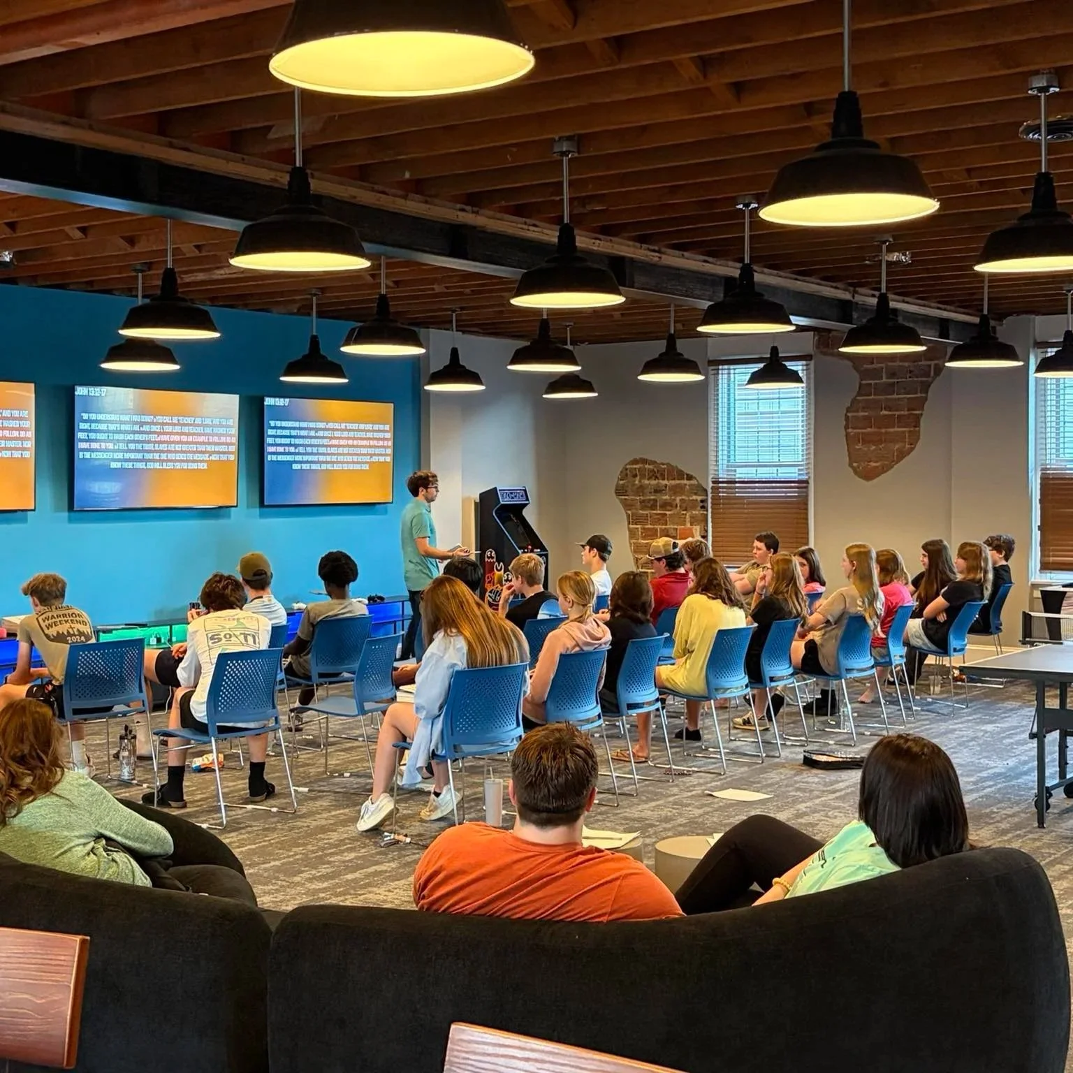 A group of young people attending a presentation or seminar in a modern, industrial-style room with brick walls and wooden ceiling. A man is standing at the front, speaking, while the audience sits on blue chairs and listens attentively.