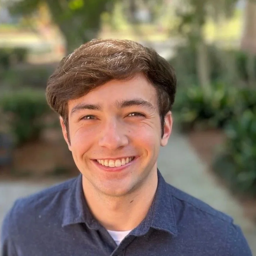 Smiling young man with brown hair, wearing a navy blue shirt, outdoors with greenery in the background.