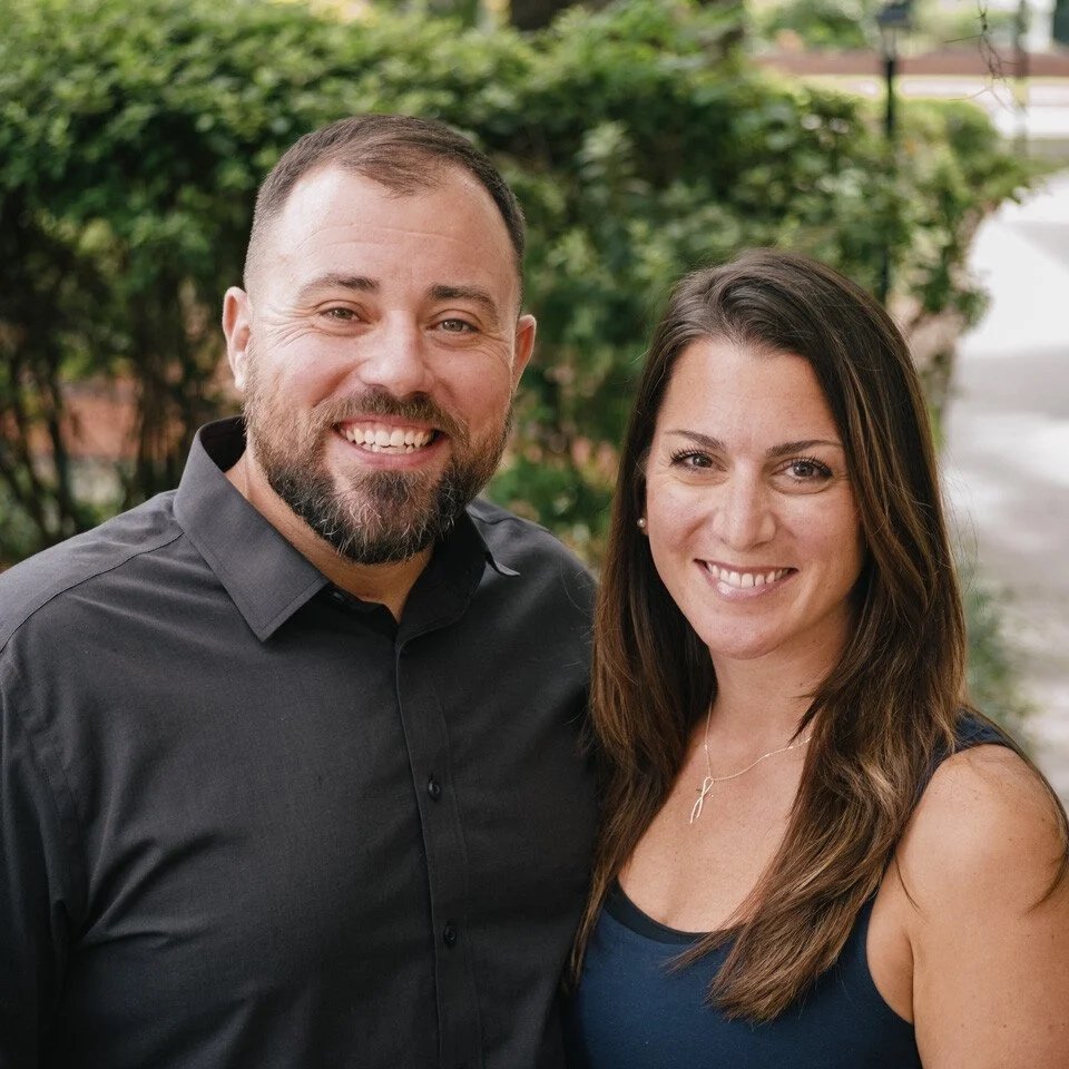 A smiling man with a beard and short hair, and a smiling woman with long brown hair, standing outdoors with greenery in the background.