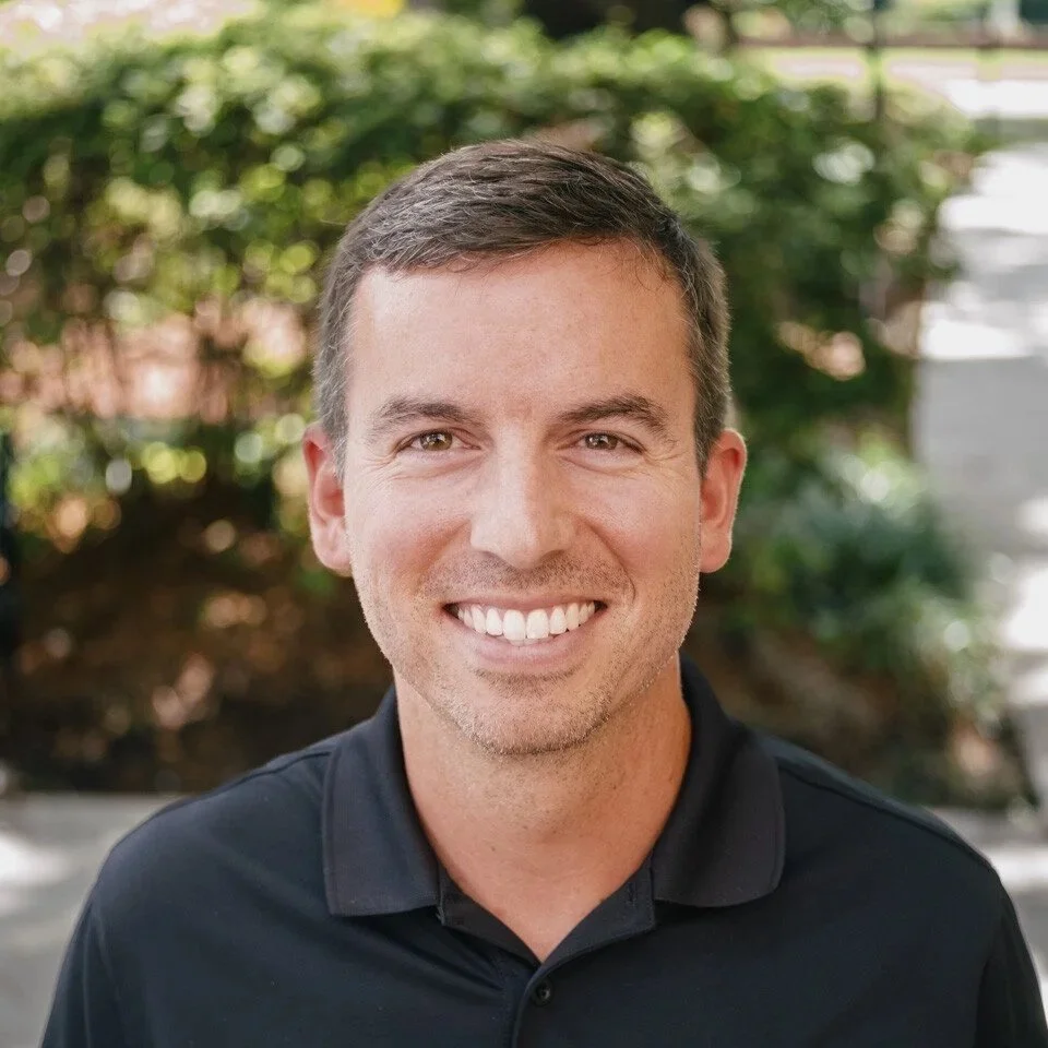 A smiling man with short brown hair wearing a black polo shirt outdoors, with green foliage and trees in the background.