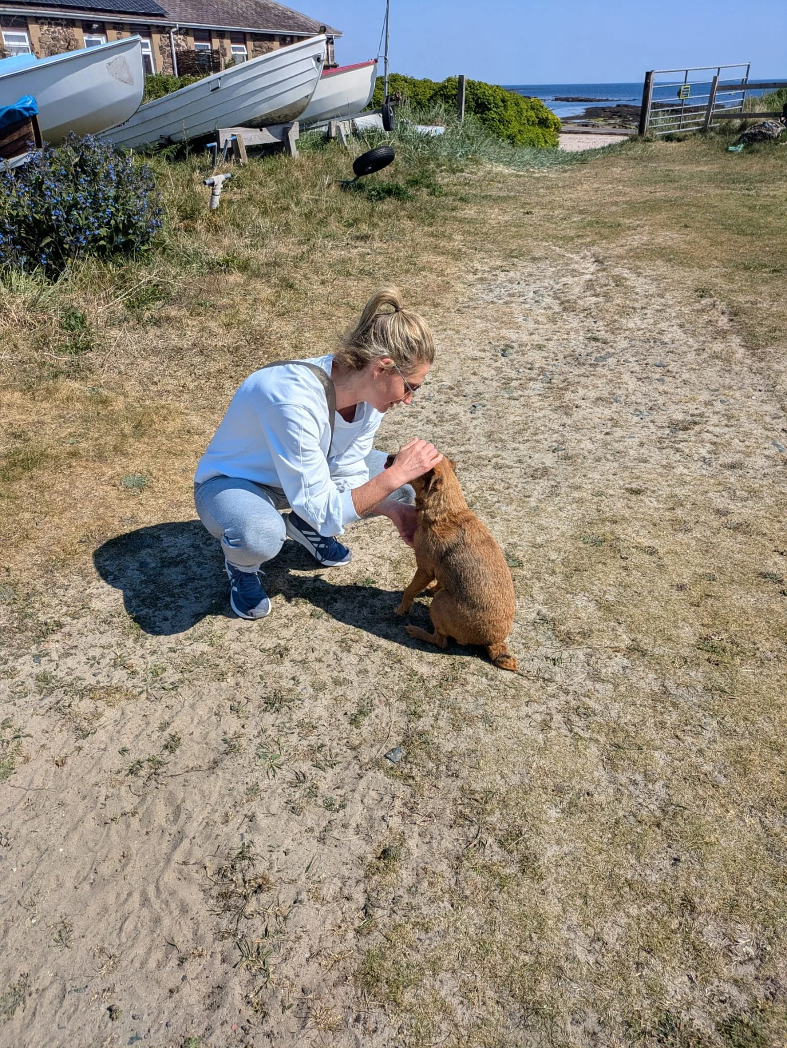 A woman with blonde hair in a ponytail, wearing a white jacket and gray pants, squatting and petting a brown dog on a dirt path near the coast. In the background, there are boats on supports, green bushes, a fence, and the ocean under a blue sky.