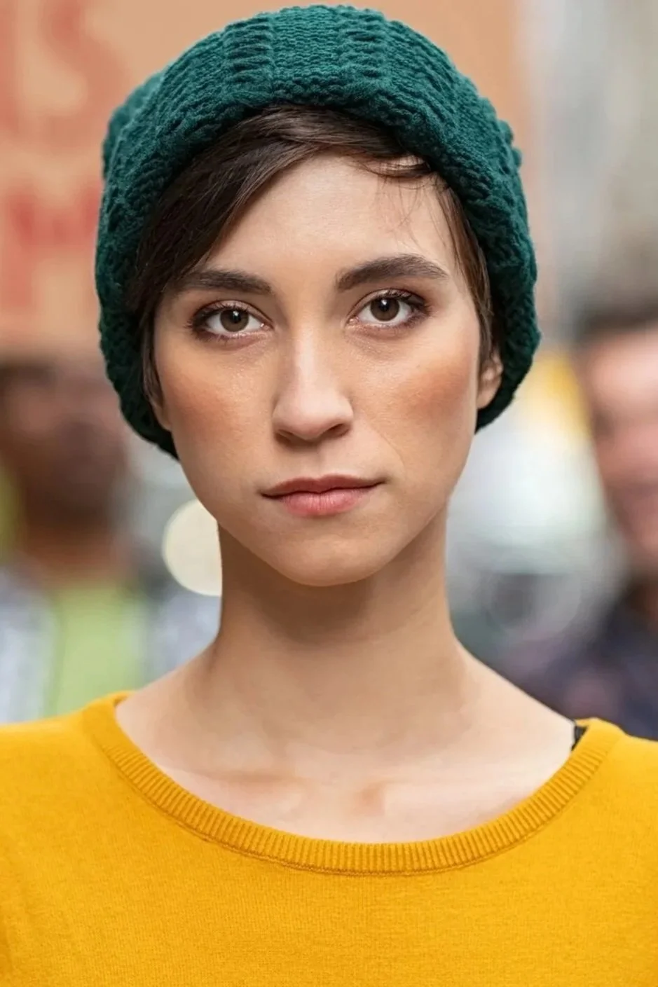 A woman with short brown hair, wearing a green knit hat and a yellow sweater, looking directly at the camera with a neutral expression. Blurred background with people.