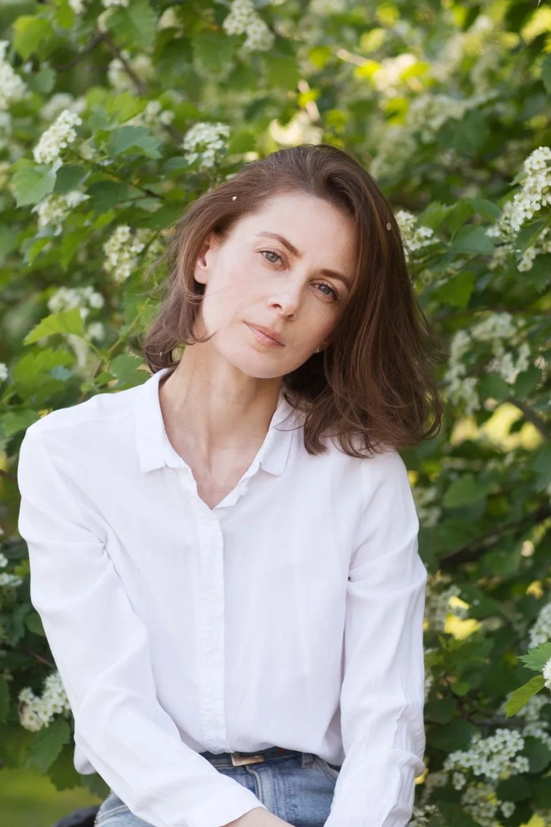 A woman with shoulder-length brown hair and light skin, wearing a white shirt, sitting outdoors among green foliage and white flowers.