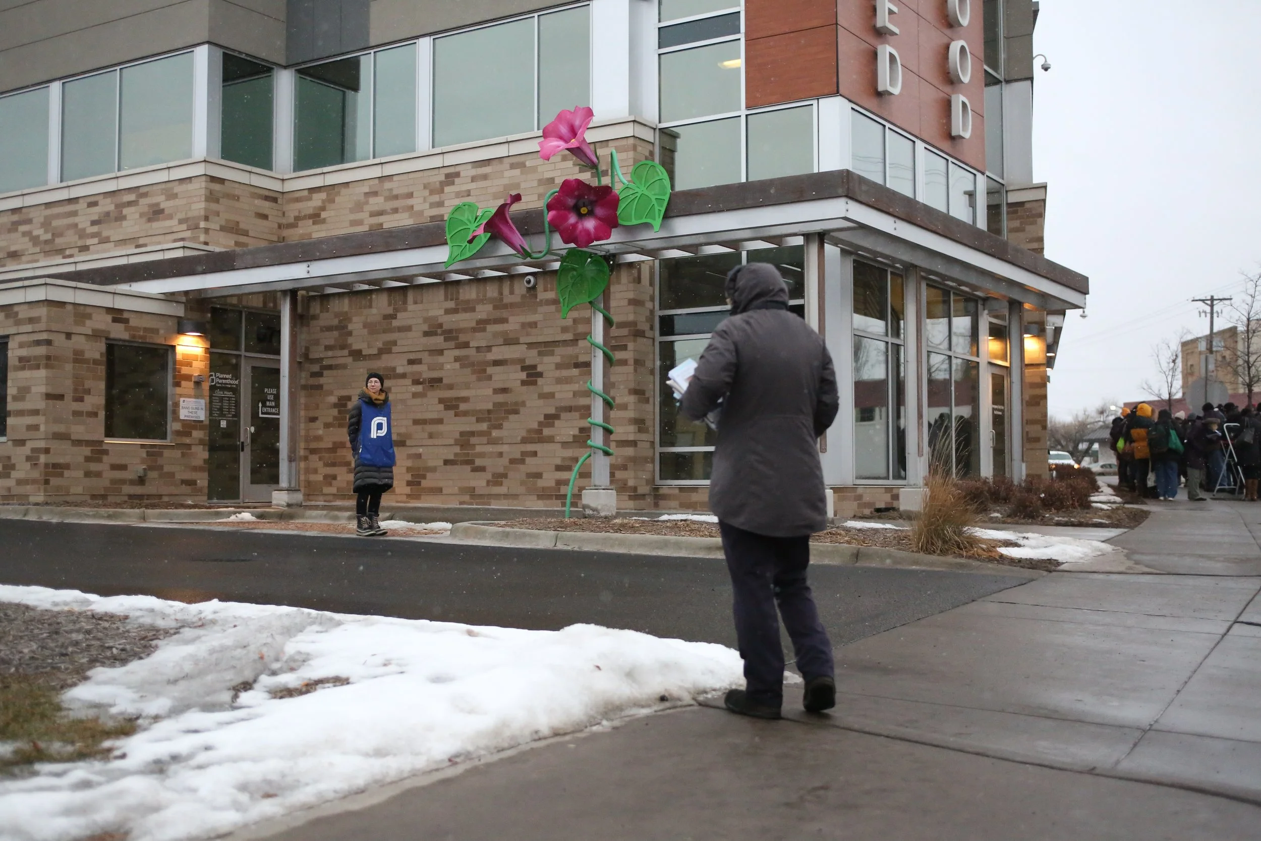 People waiting outside a brick building with a colorful flower decoration on the corner of the building.