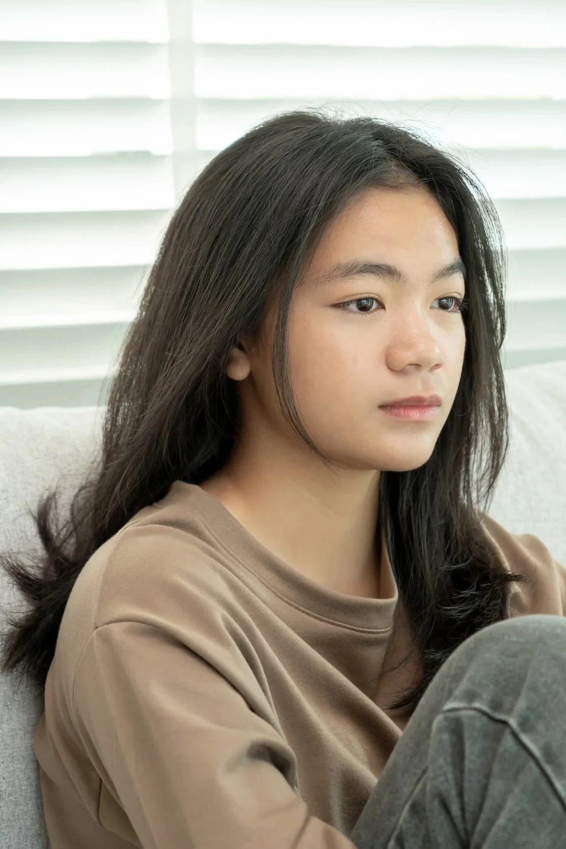 A young woman with long dark hair sitting on a couch, wearing a beige top and looking at a device in her hands, with white window blinds in the background.