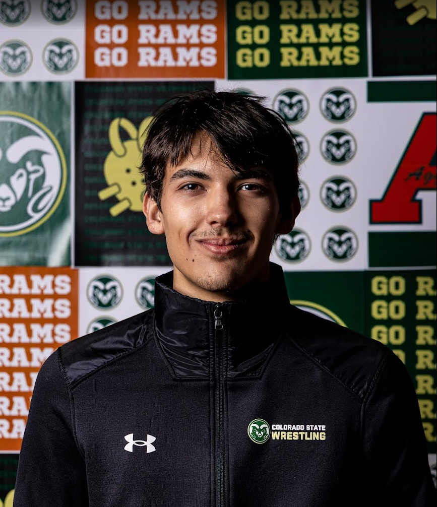 Young man in black Colorado State Wrestling jacket standing in front of a backdrop with green, yellow, orange, and red Rams logos and slogans.