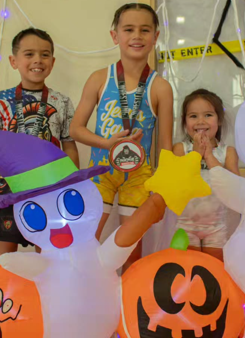 Three children standing behind Halloween farm decorations, including a paper ghost with a witch hat and a pumpkin. The children are wearing medals, smiling, and holding a star-shaped object.