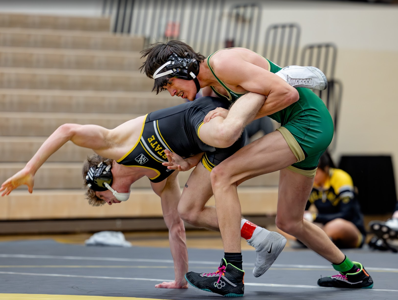 Two male wrestlers in a match on a grey mat, wearing singlets and headgear, with one in black and yellow and the other in green and beige, in a gymnasium with wooden bleachers.