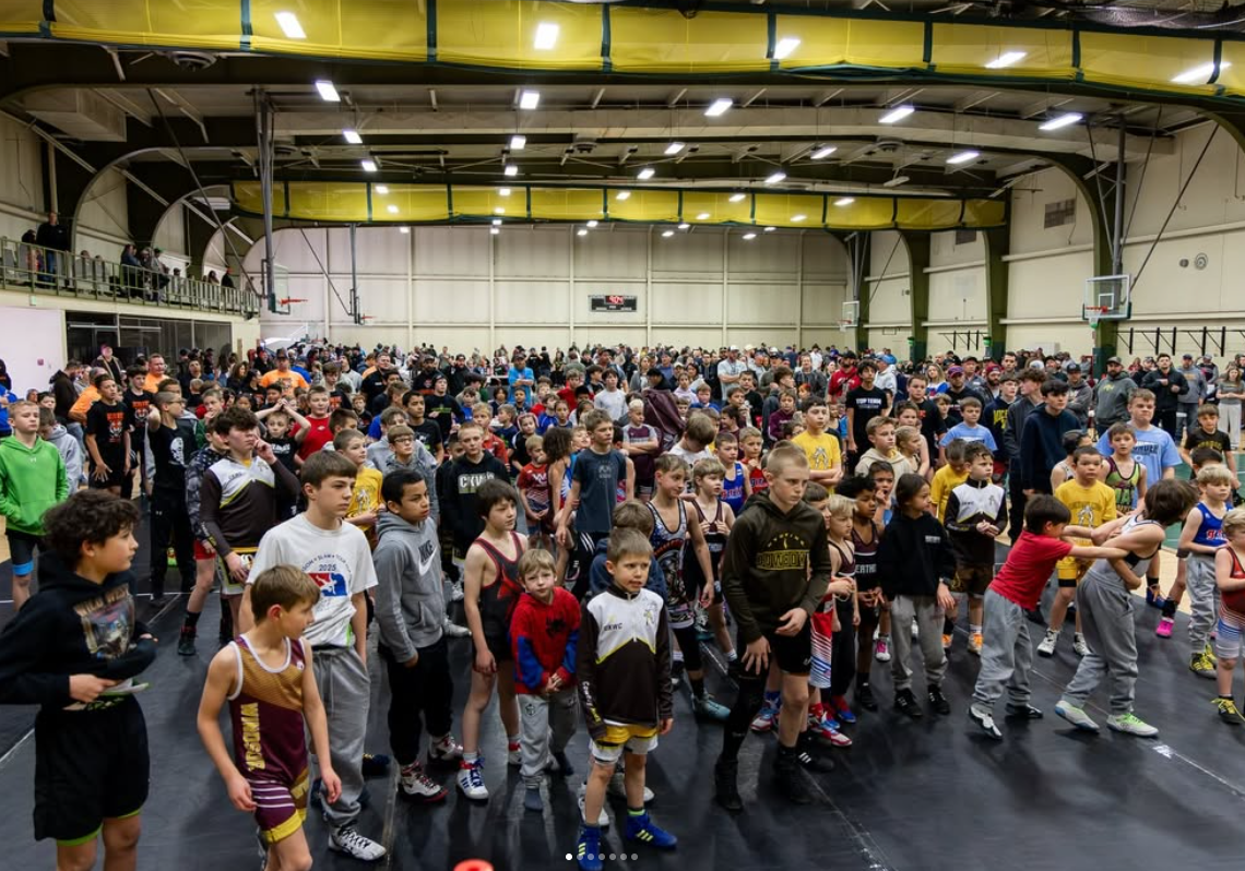 A large indoor wrestling tournament with many young wrestlers in colorful uniforms, some stretching or warming up, in a gymnasium with spectators watching from a balcony.